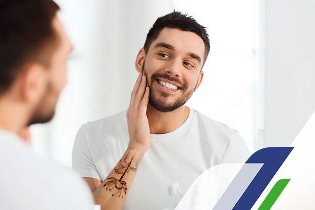 Homem jovem de barba e com o braço tatuado se olhando no espelho com um sorriso no rosto