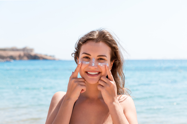 Mulher sorrindo e aplicando protetor solar no rosto em dia ensolarado na praia, reforçando a importância da proteção solar.